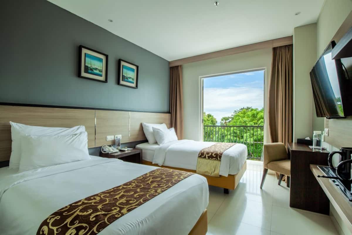 Ein Hotelzimmer mit zwei Betten, einem Schreibtisch, einem Fernseher und einem Balkon mit Blick auf Grünflächen.