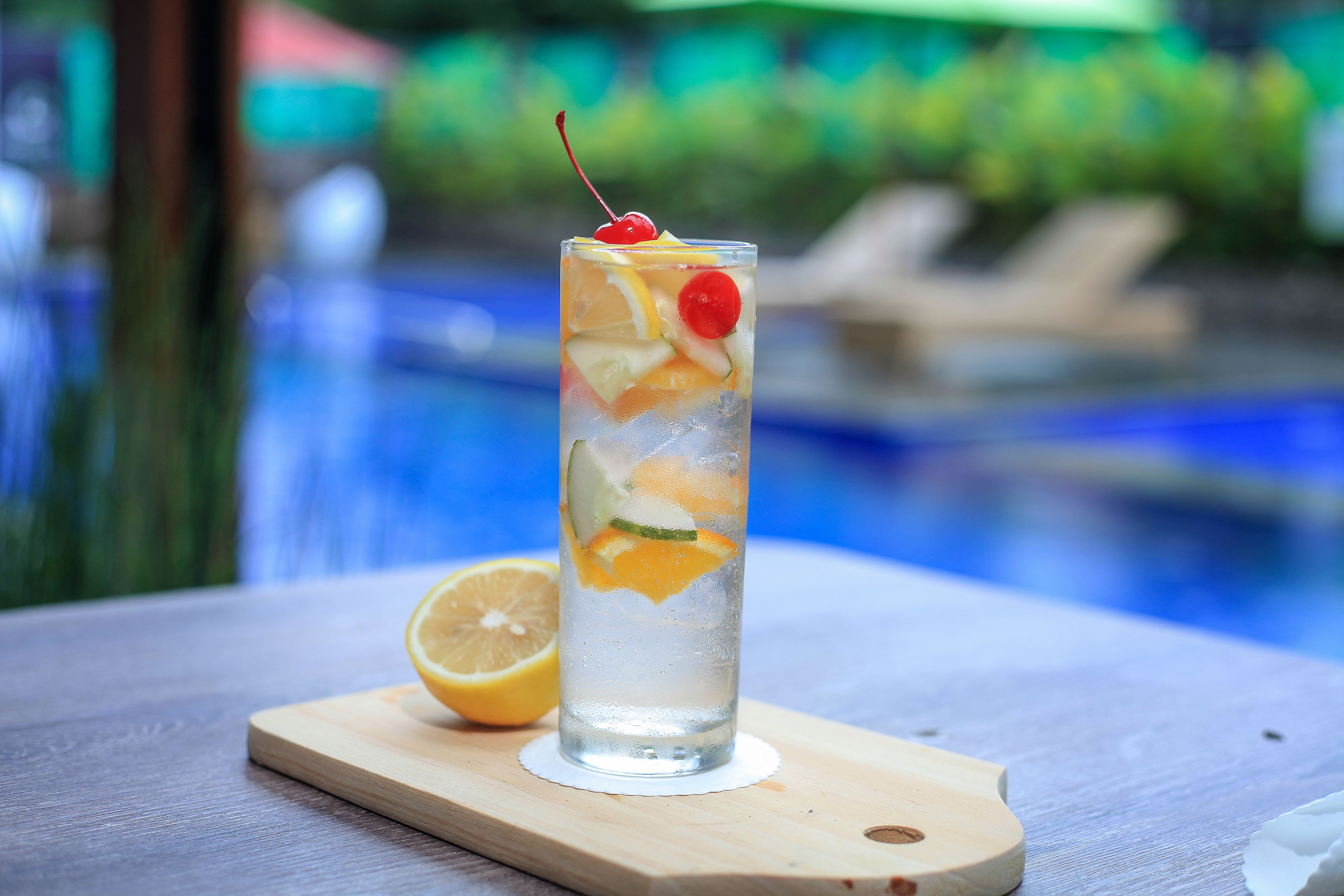 A tall glass with a cherry on top, containing ice and fruit slices, placed on a wooden board next to a half lemon.