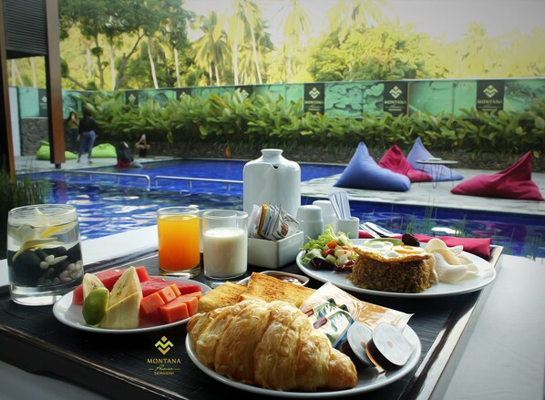 A breakfast tray with croissants, fruits, and beverages, placed on a table with a pool view in the background. - Montana Premier Senggigi (Senggigi)