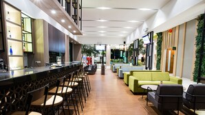 Un moderno bar con sillas de madera y una barra, un área de descanso con sofás verdes, y una pantalla de televisión.