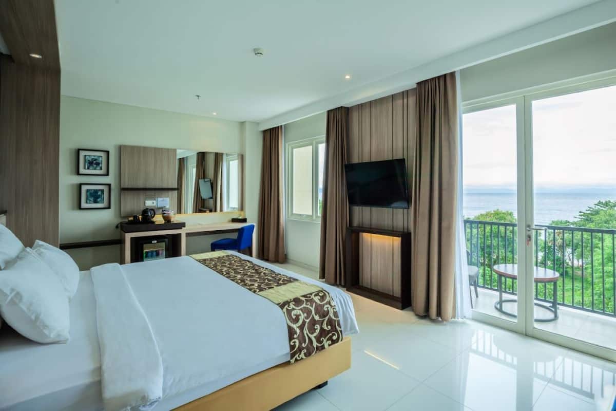 Ein Hotelzimmer mit einem großen Bett, einem Schreibtisch, einem Fernseher und einem Balkon mit Meerblick.