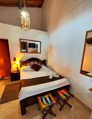 A neatly made bed with a headboard, a bedside table with a lamp, a mirror above the bed, and a colorful stool in front of the bed. - Hostel Santander Aleman Terrace Vista (San Gil)