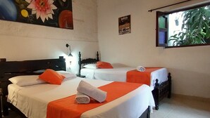 Quarto de hotel com duas camas, cada uma com roupa de cama branca e detalhes em laranja, um quadro na parede, um cartaz e uma janela com vista para o verde.
