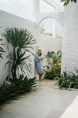 A person standing in a white-walled indoor space with large plants and a tiled floor.