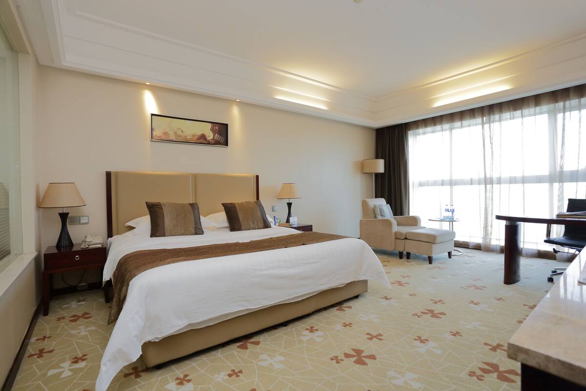 Une chambre d’hôtel avec un grand lit, deux tables de chevet avec des lampes, un bureau, une chaise et une grande fenêtre avec des rideaux.