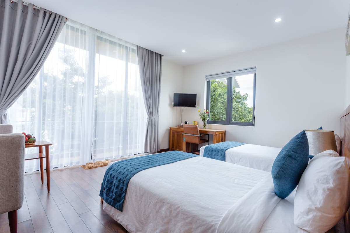 Une chambre d’hôtel avec deux lits, un bureau, une chaise, une télévision et de grandes fenêtres avec des rideaux.