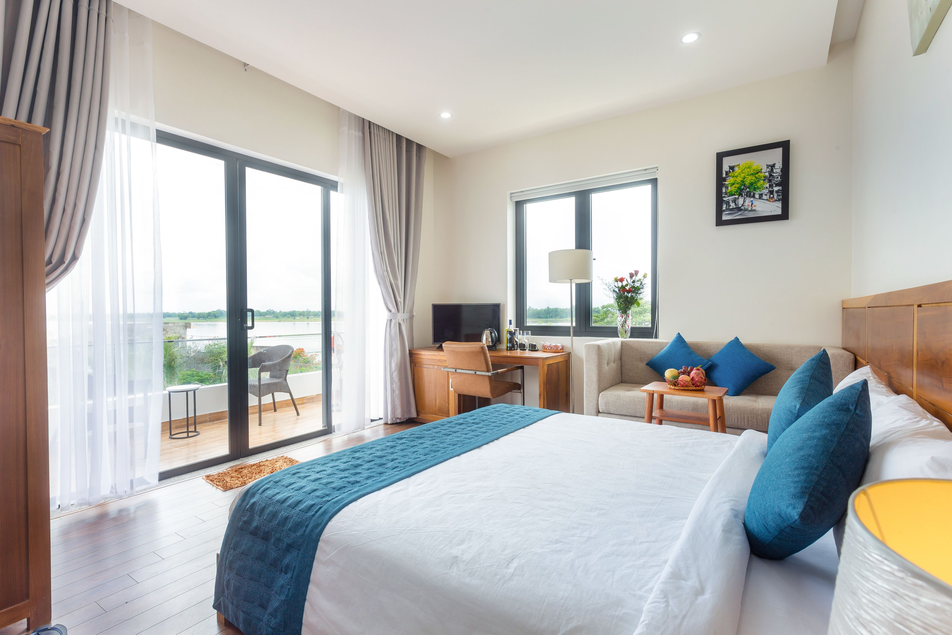 Hotelkamer met een groot bed, een bureau, een stoel en een bank. Er is een balkon met uitzicht op groen.