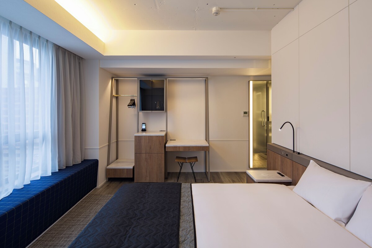 A modern hotel room with a large bed, a desk, a small seating area, and a bathroom with a mirror.