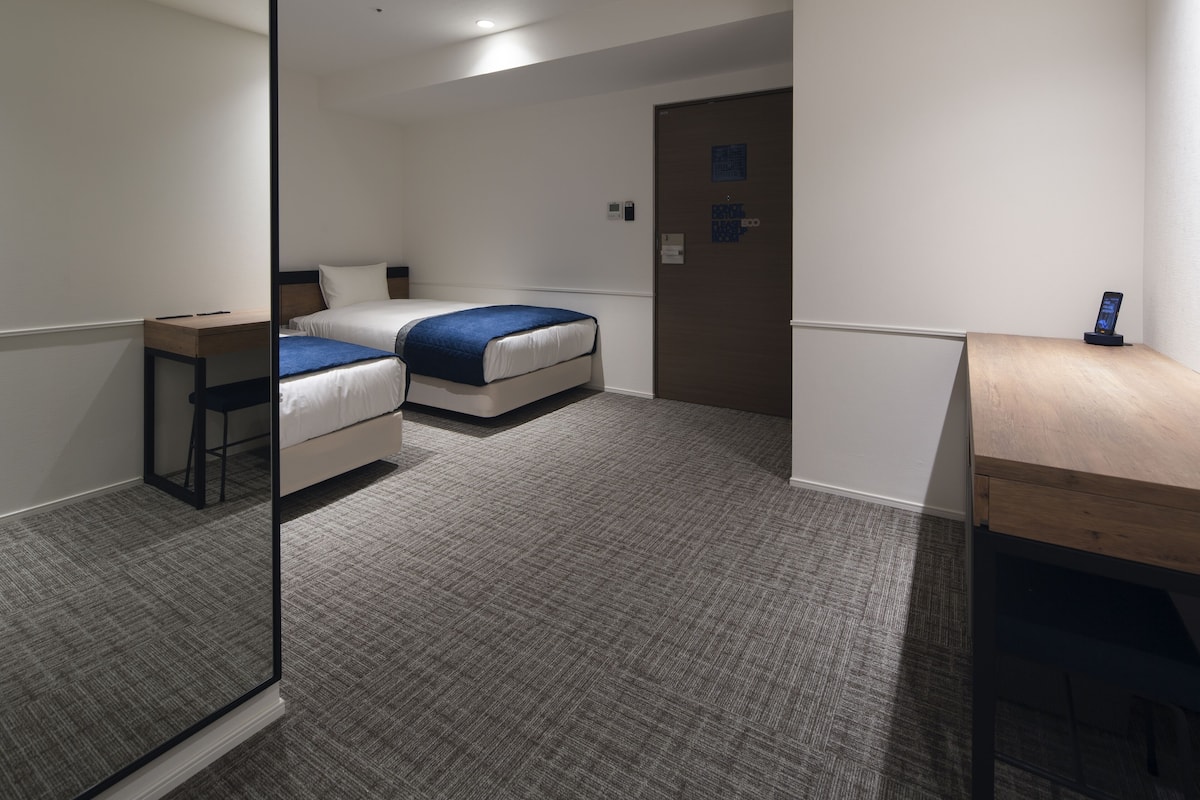 A hotel room with two single beds, a desk, a mirror, and a door with a lock.