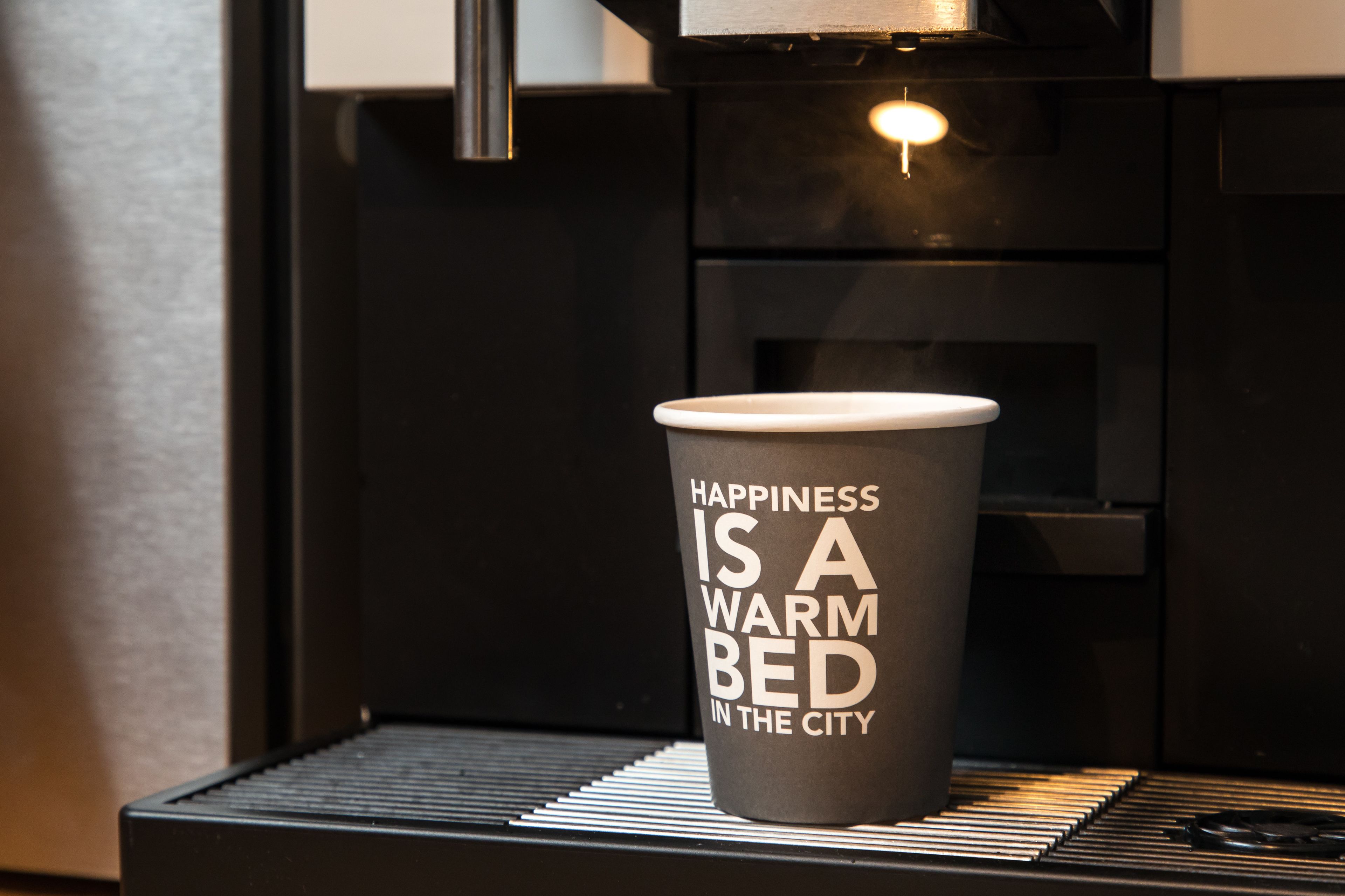 A cup with the text "Happiness is a warm bed in the city" placed on a tray in front of a coffee machine.