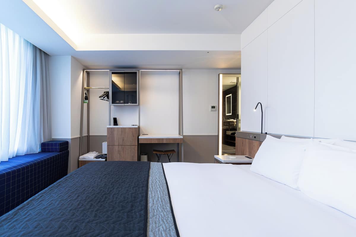 A modern hotel room with a large bed, a desk, a mirror, and a wardrobe.