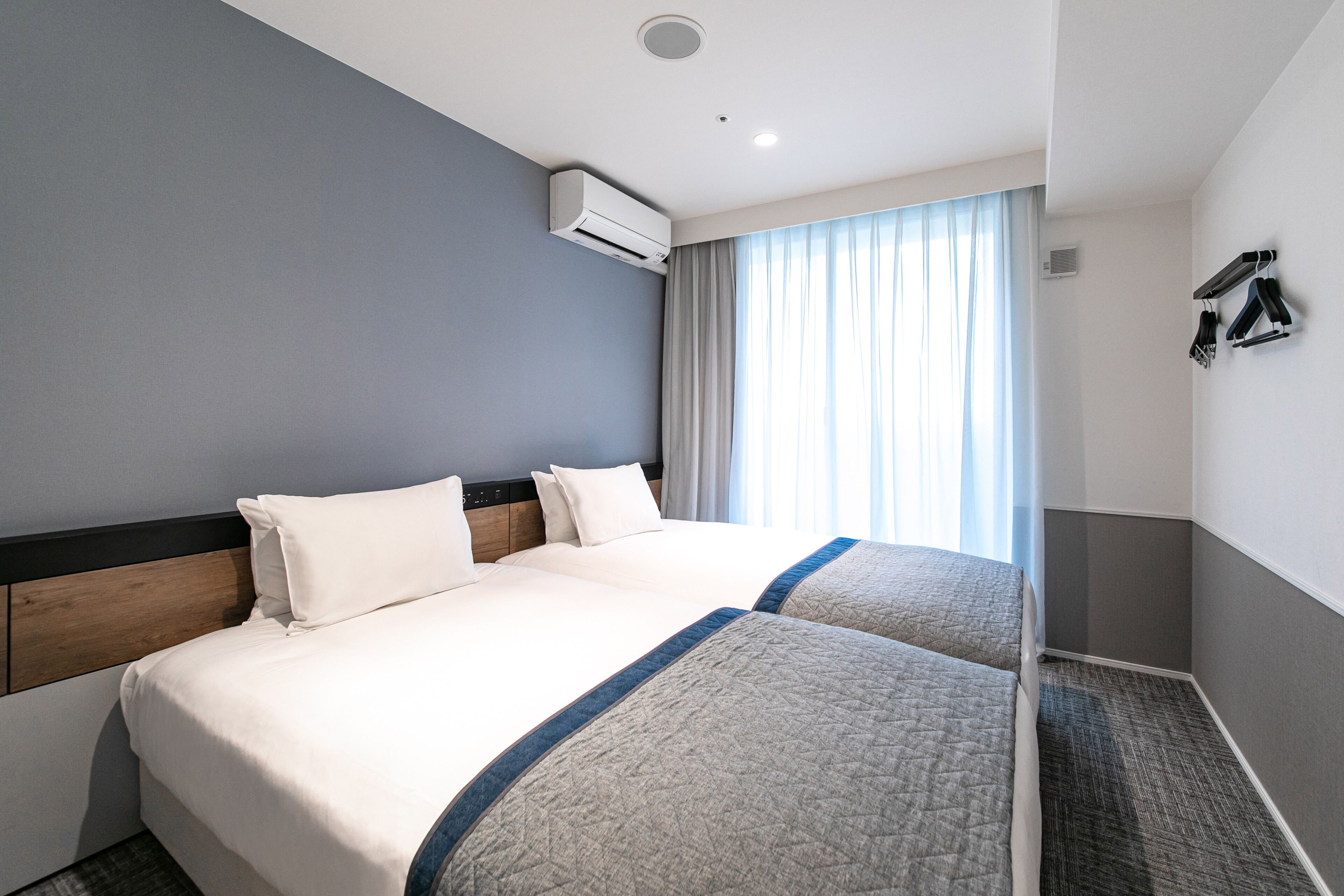 A hotel room with two beds, a grey wall, a window with curtains, and an air conditioning unit.