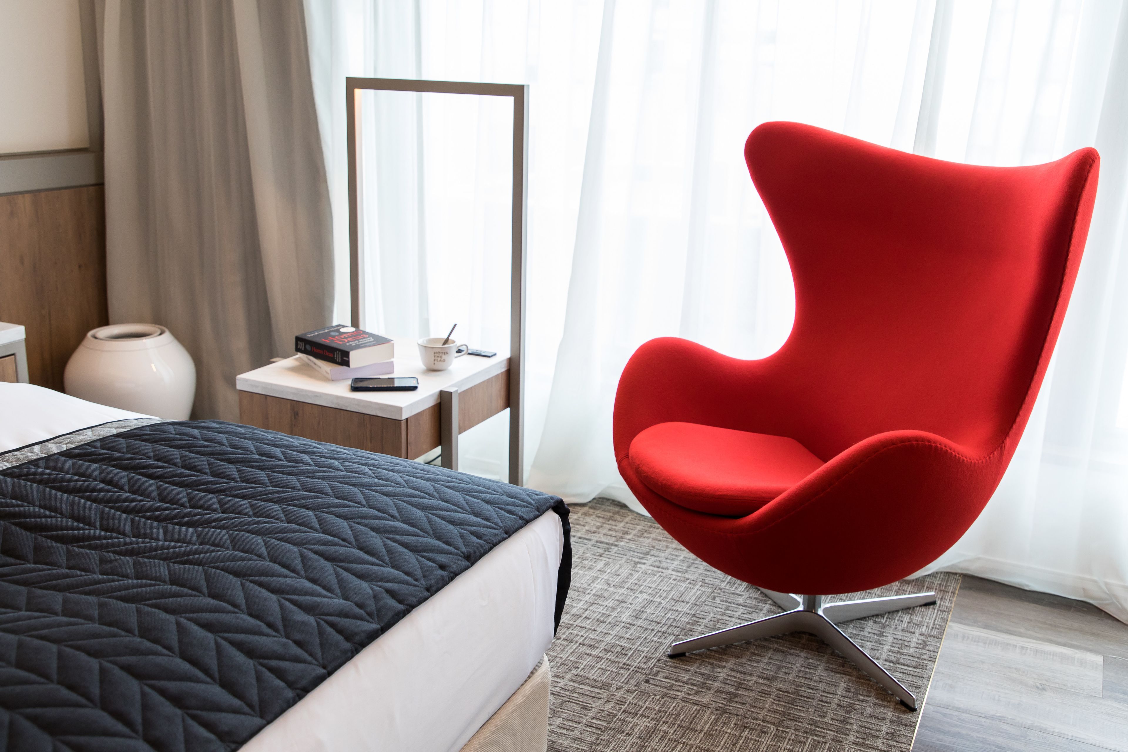 A hotel room with a bed, a red chair, a desk with a mirror, and a window with curtains.