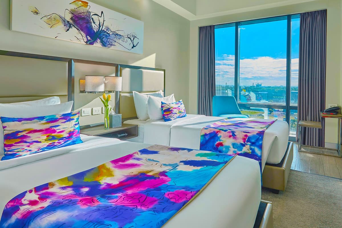A hotel room with two beds, a large window with a city view, a desk with a lamp, and a painting on the wall.