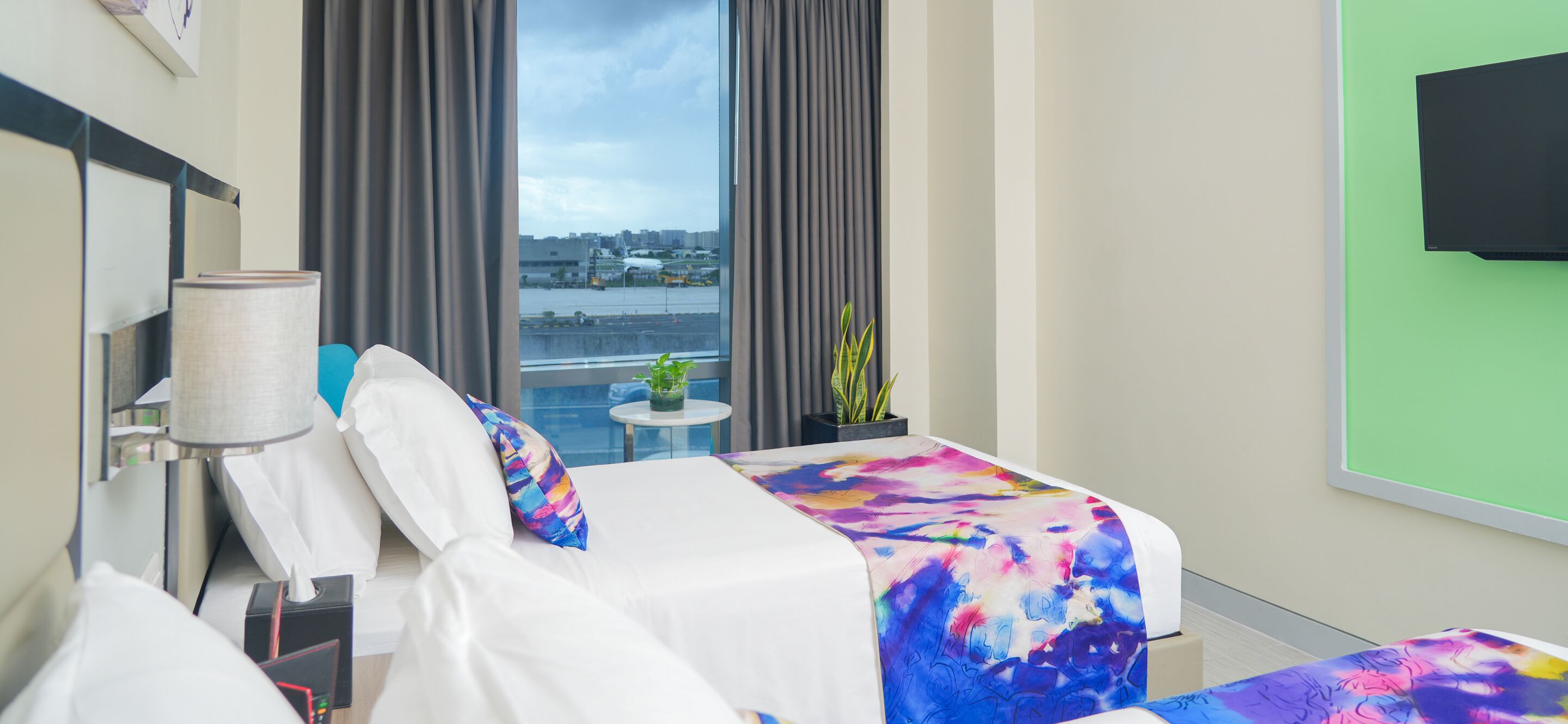 Une chambre d’hôtel avec un lit, une table de chevet, une lampe, une petite table avec une plante et une vue sur la ville grâce à de grandes fenêtres.