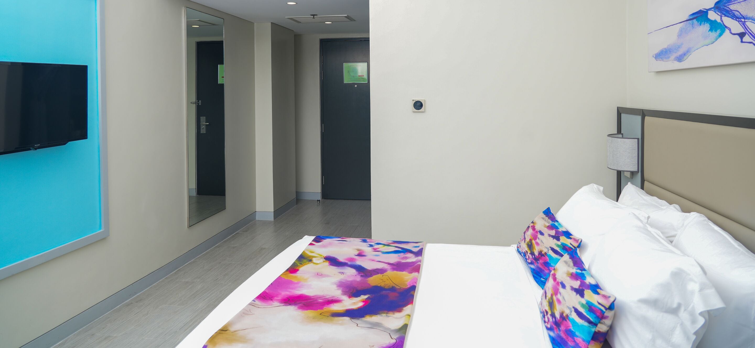Habitación de hotel con una cama, un televisor montado en la pared, una puerta y un cuadro.
