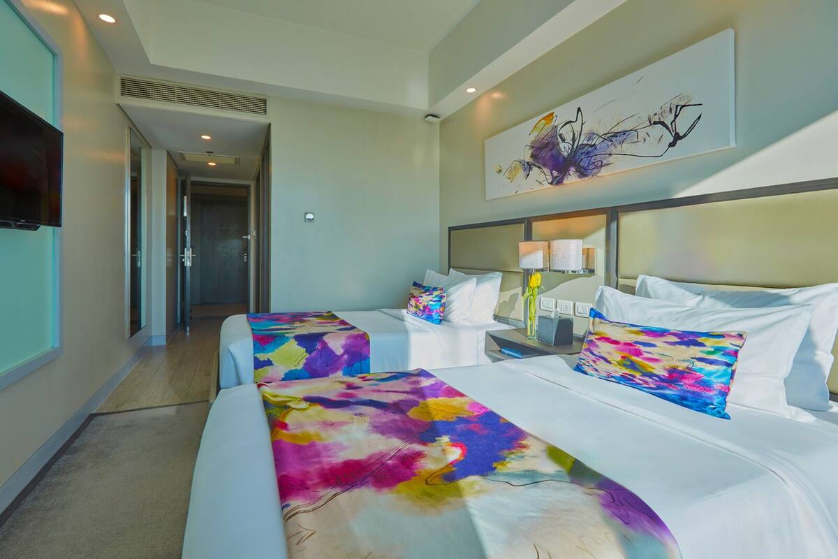 A hotel room with two beds, a large abstract painting on the wall, and a television mounted on the wall.