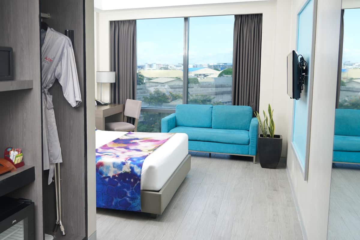 A hotel room with a bed, a blue sofa, a TV, a window with a city view, and a wardrobe.
