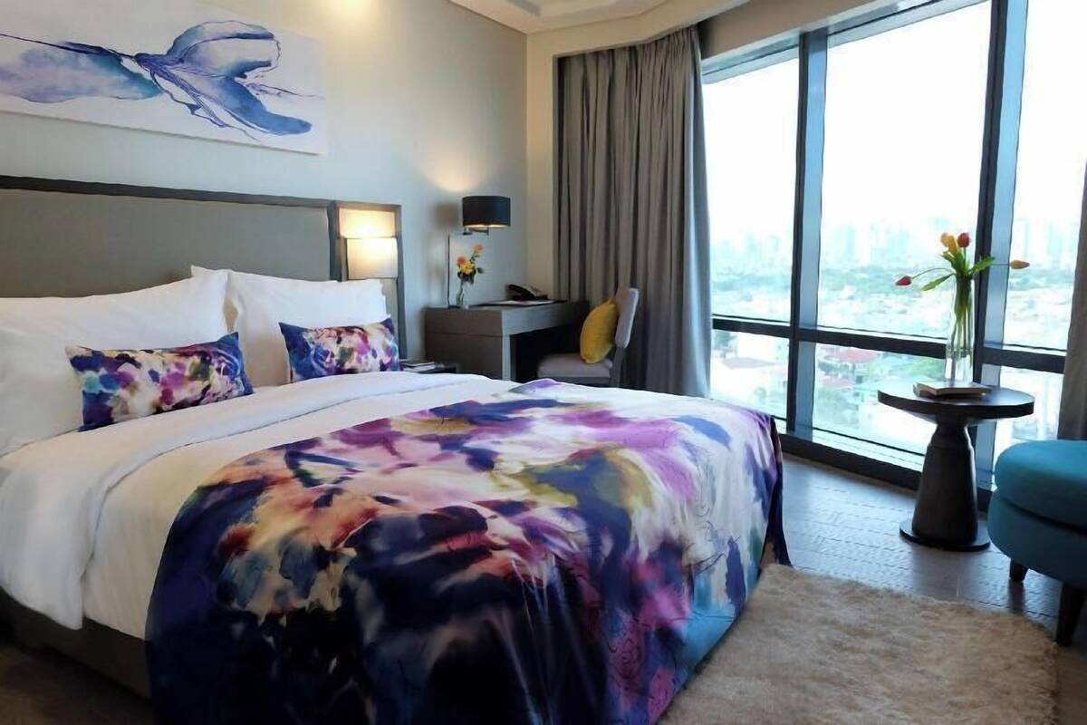 A hotel room with a large bed, a desk, a chair, a window with a city view, and a painting on the wall.
