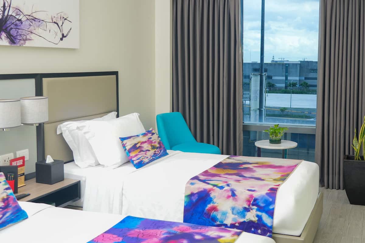 A hotel room with two beds, a nightstand, a small table, and a window with curtains.