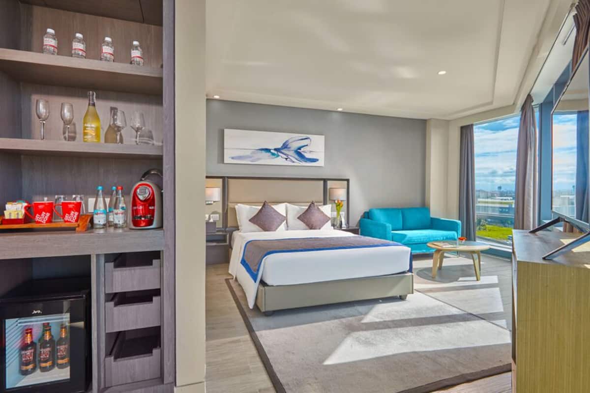 A modern hotel room with a bed, a blue sofa, a coffee table, and a minibar.