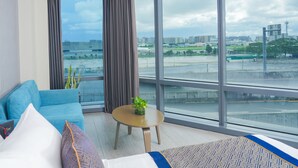 Una habitación de hotel moderna con un ventanal grande, un sofá azul, una mesita redonda con una planta y una cama con ropa de cama blanca y azul.
