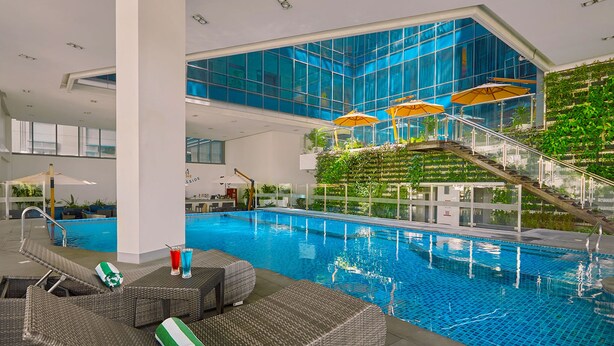 Una piscina interior con sillones de descanso y sombrillas, rodeada de paredes de vidrio y una escalera con vegetación.