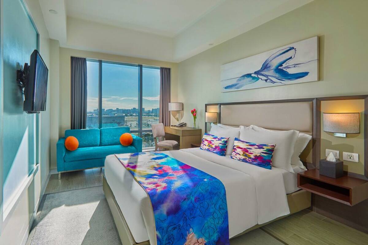 A hotel room with a large bed, a blue sofa, a city view, and a painting on the wall.