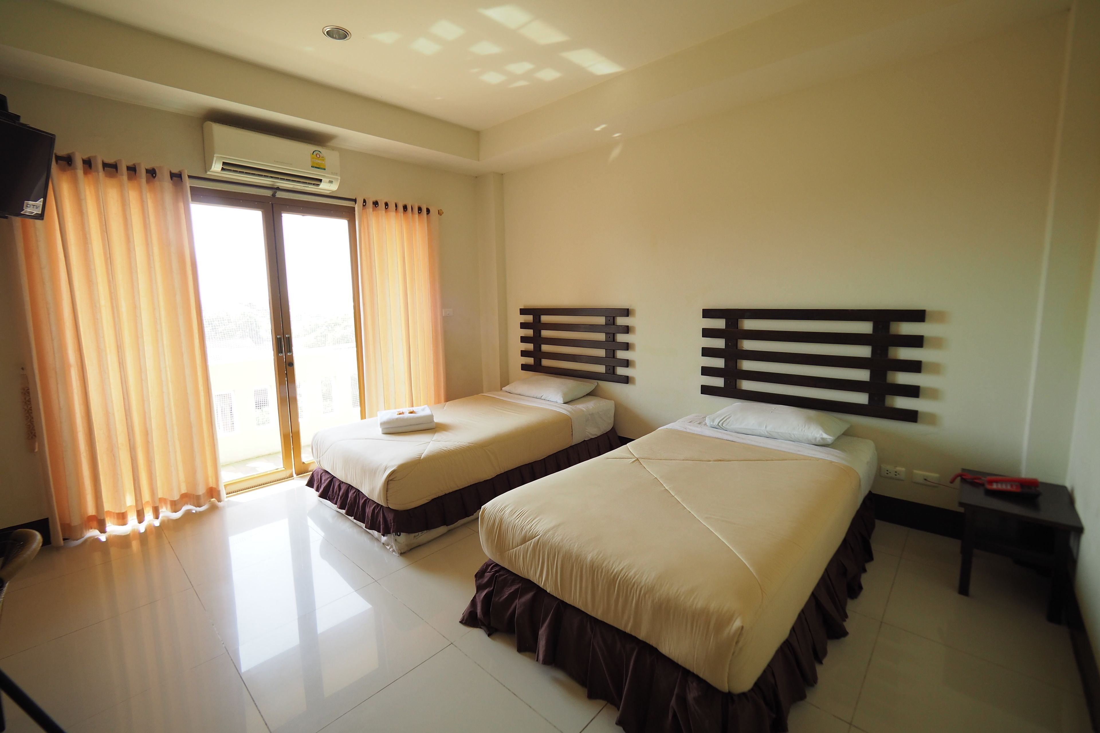 A hotel room with two beds, a ceiling fan, an air conditioning unit, a window with curtains, and a small table with a red object on it.