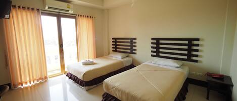 A hotel room with two beds, a ceiling fan, an air conditioning unit, a window with curtains, and a small table with a red object on it.