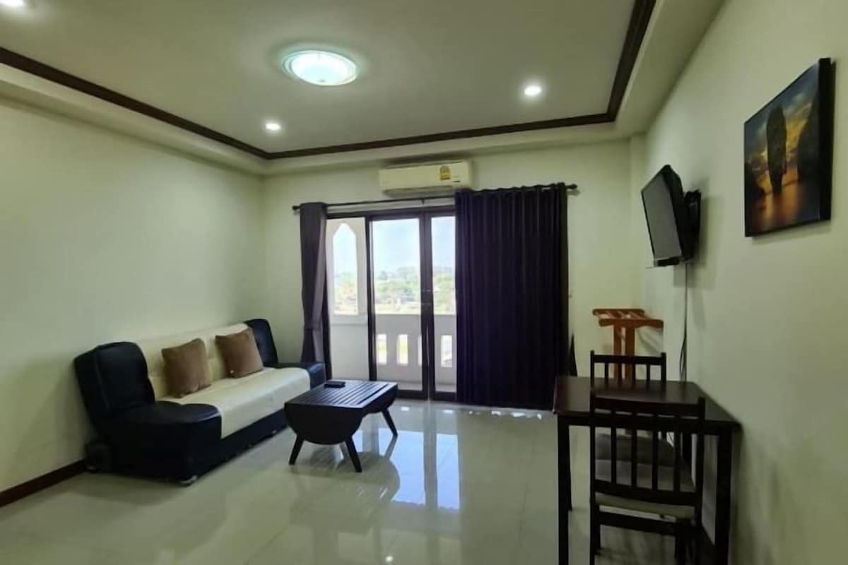 Habitación de hotel con sofá, una pequeña mesa, televisor montado en la pared y balcón con cortinas.