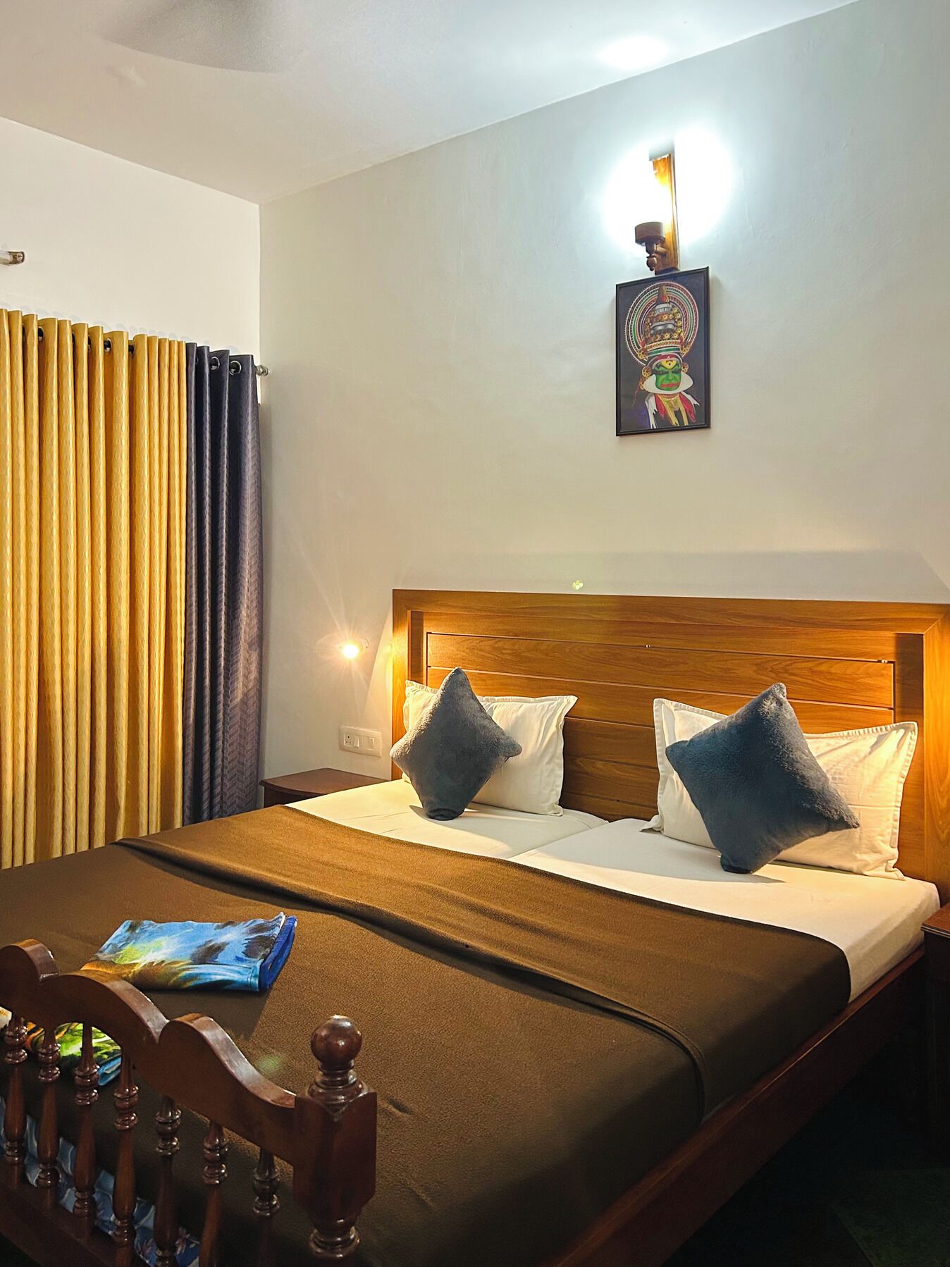 A hotel room with a wooden headboard, two pillows, a bed with a brown bedspread, a nightstand with a lamp, and a painting on the wall.