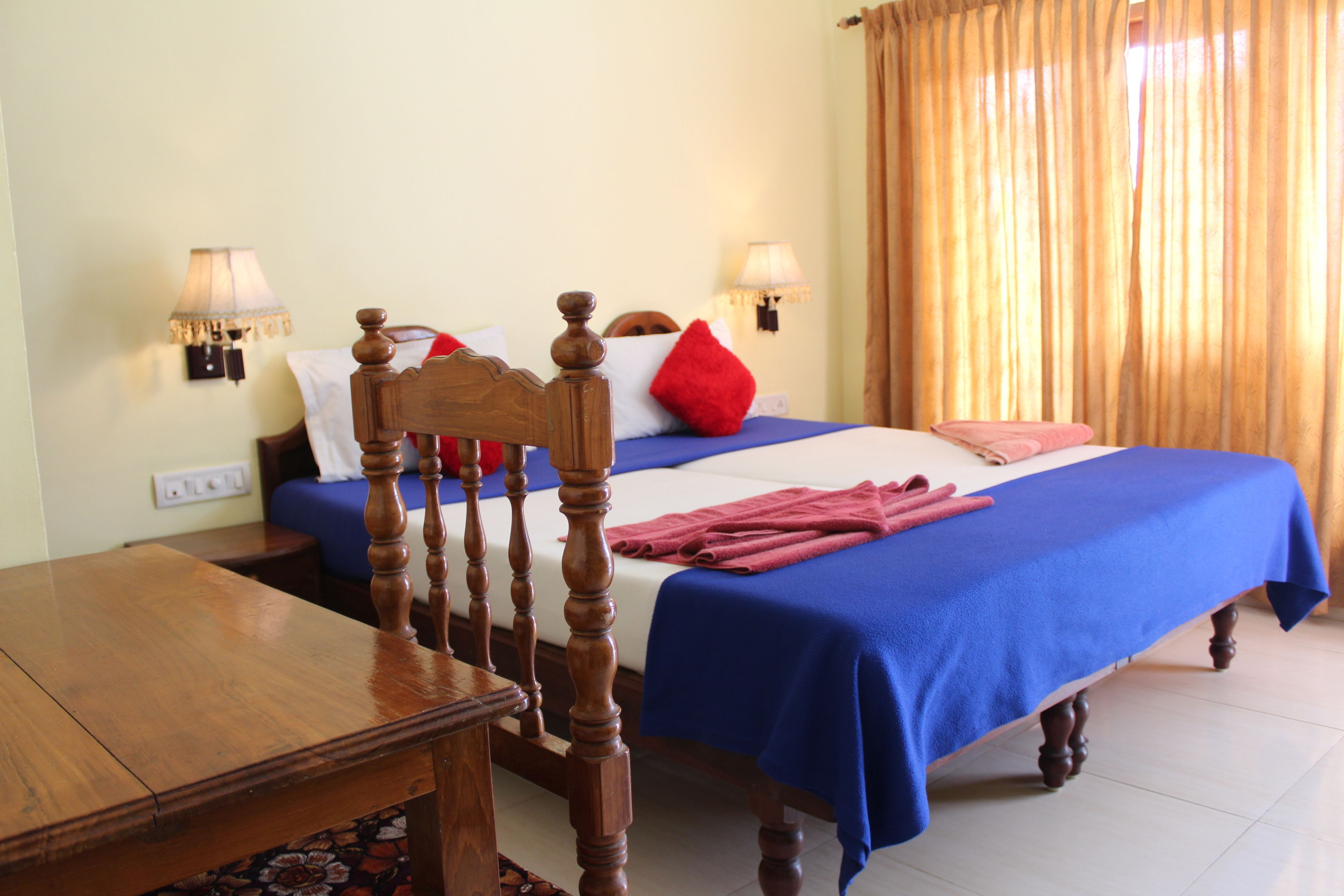 A hotel room with a wooden bed, blue bedspread, red and white pillows, two bedside lamps, and a wooden nightstand.