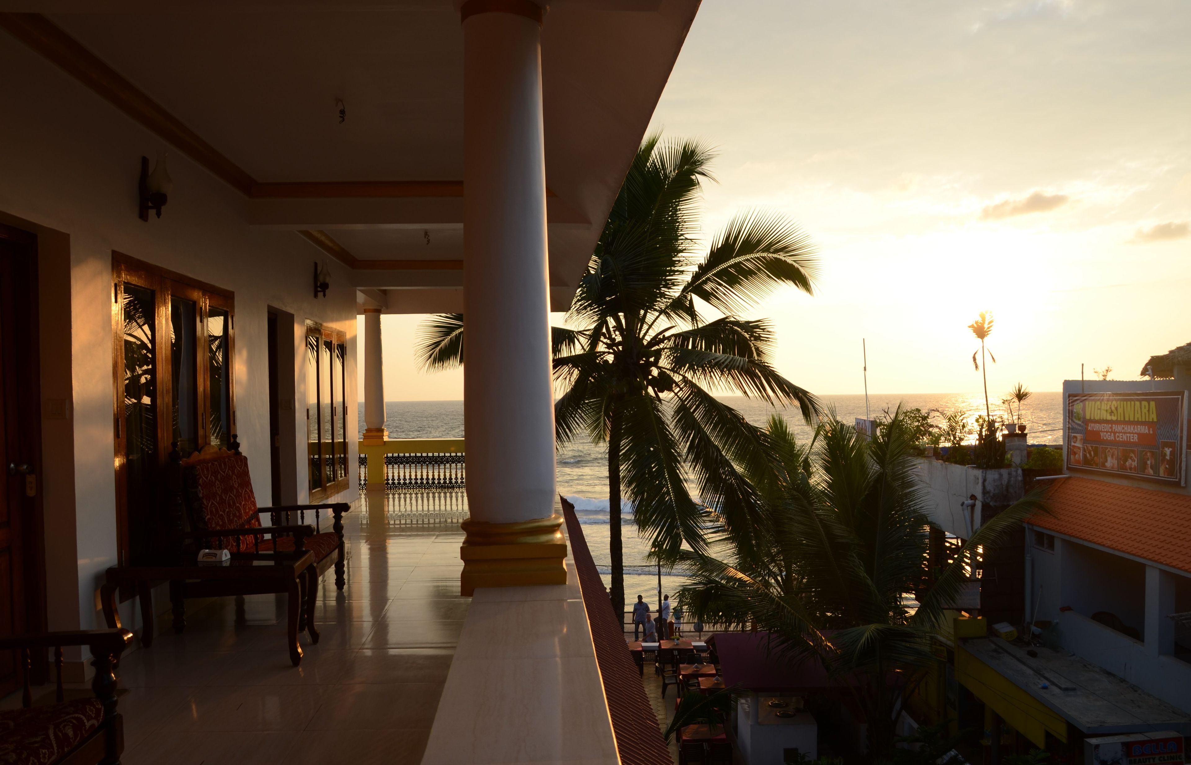 A hotel corridor with a view of the ocean at sunset, featuring a palm tree, a bench, and a railing.