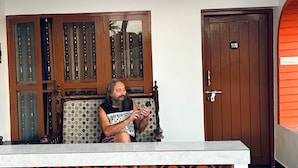 A man sitting on a patterned sofa in a room with a wooden cabinet and a door marked with the number 116.