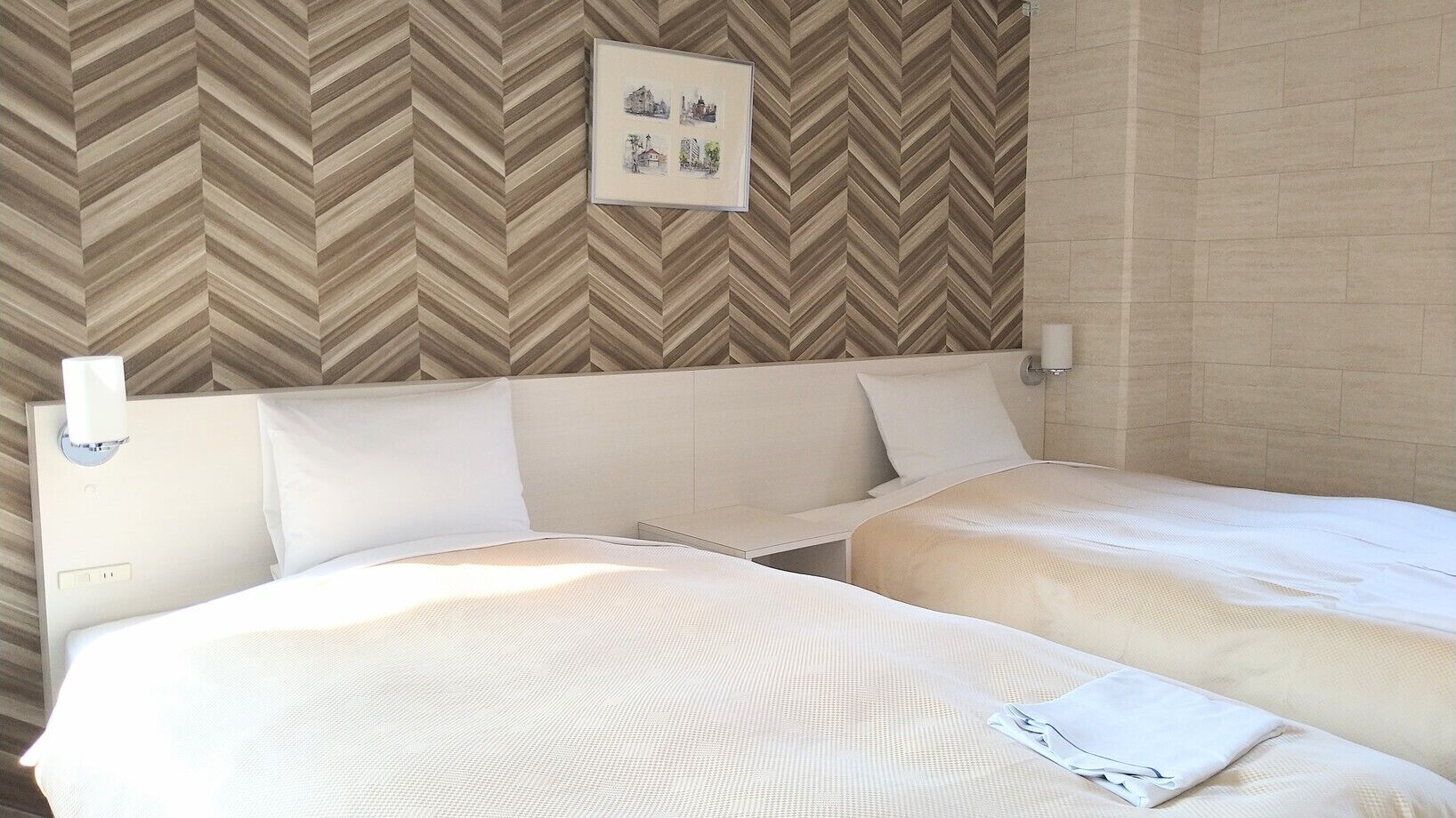 Habitación de hotel con dos camas, una pared con diseño de estampada en forma de V, un cuadro enmarcado y una lámpara de noche.