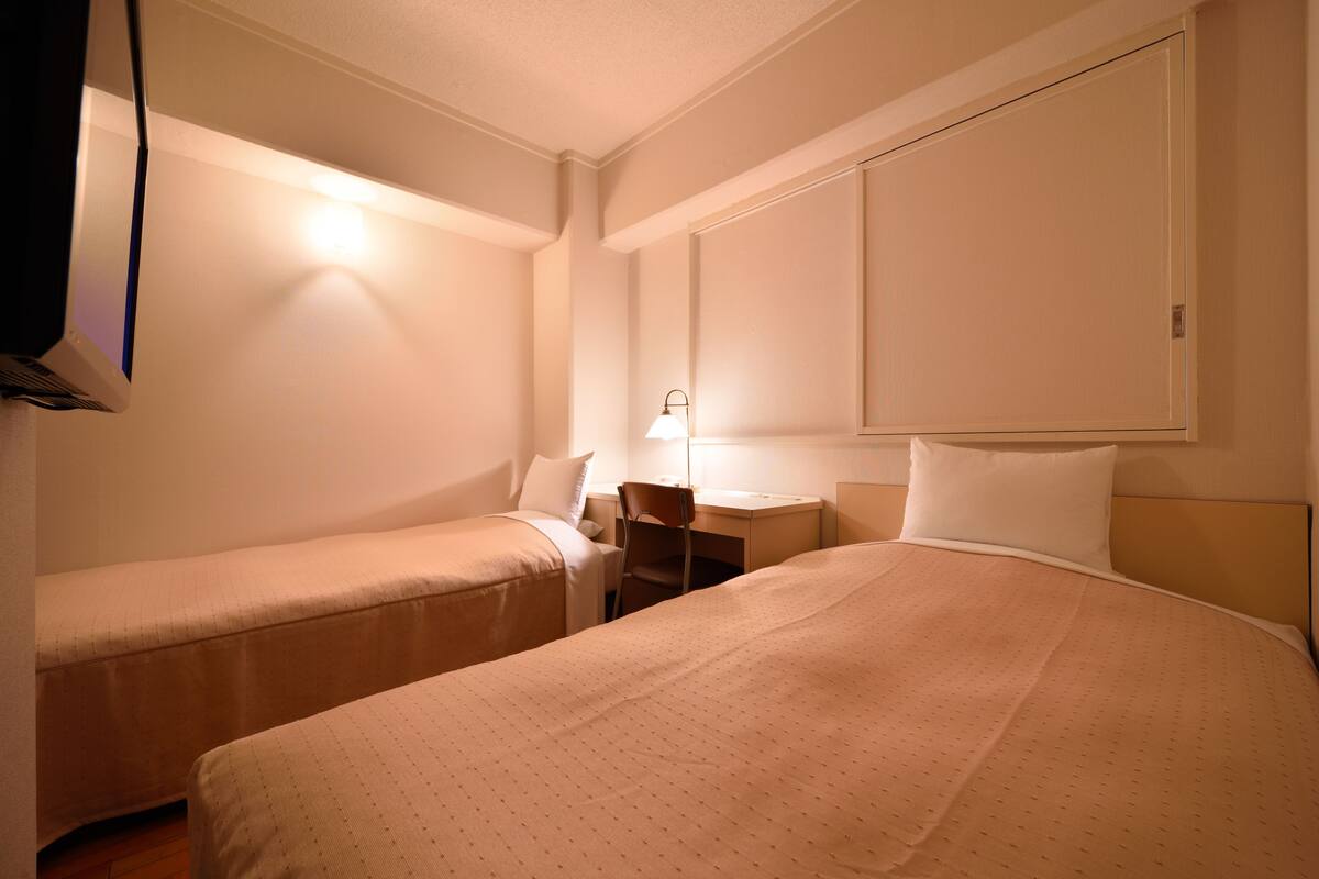 Une chambre d’hôtel avec deux lits simples, un bureau avec une chaise, un téléviseur fixé au mur et une table de chevet avec une lampe.