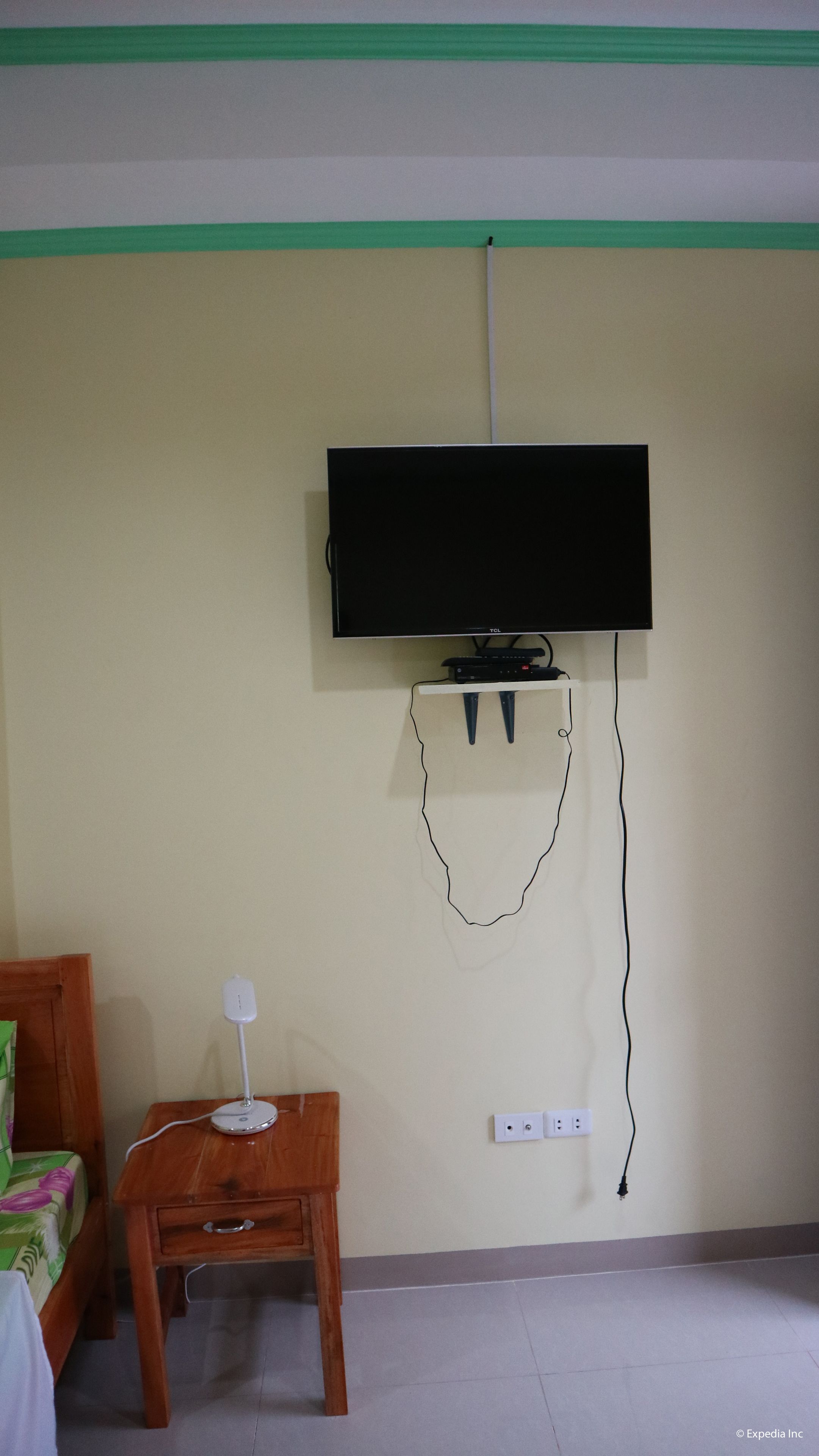 A room with a television mounted on the wall, a small wooden table with a lamp, and a bed with a green and white bedspread.
