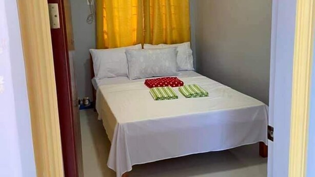 Una cama pequeña y bien hecha con sábanas blancas y dos almohadas verde a rayas, una cortina amarilla y una pared blanca con detalles azules.