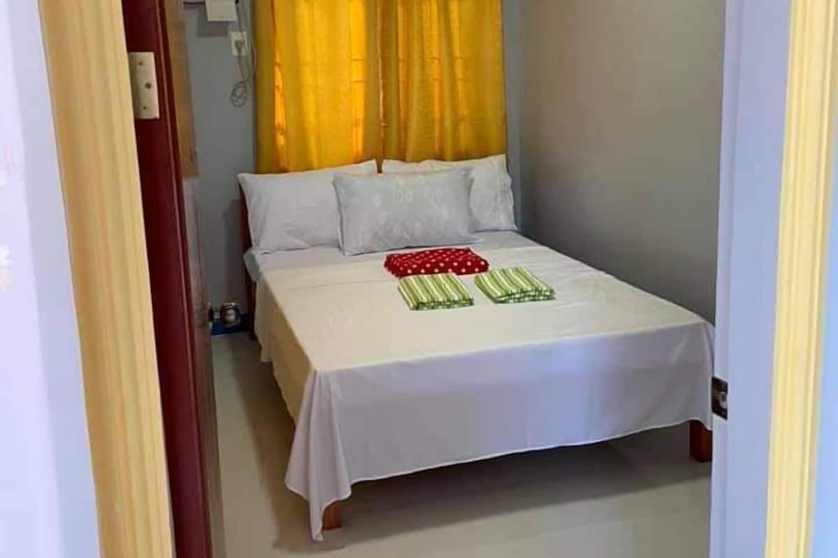 A small, neatly made bed with white linens and two green striped pillows, a yellow curtain, and a white wall with a blue trim.