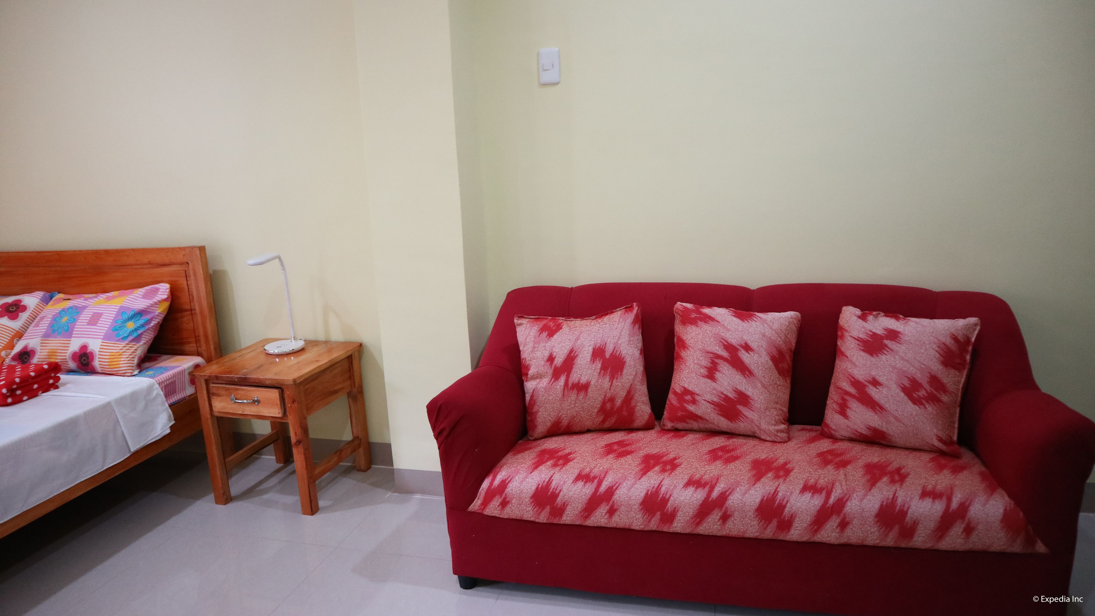 A room with a bed, a wooden nightstand, a red sofa, and a small table with a lamp.