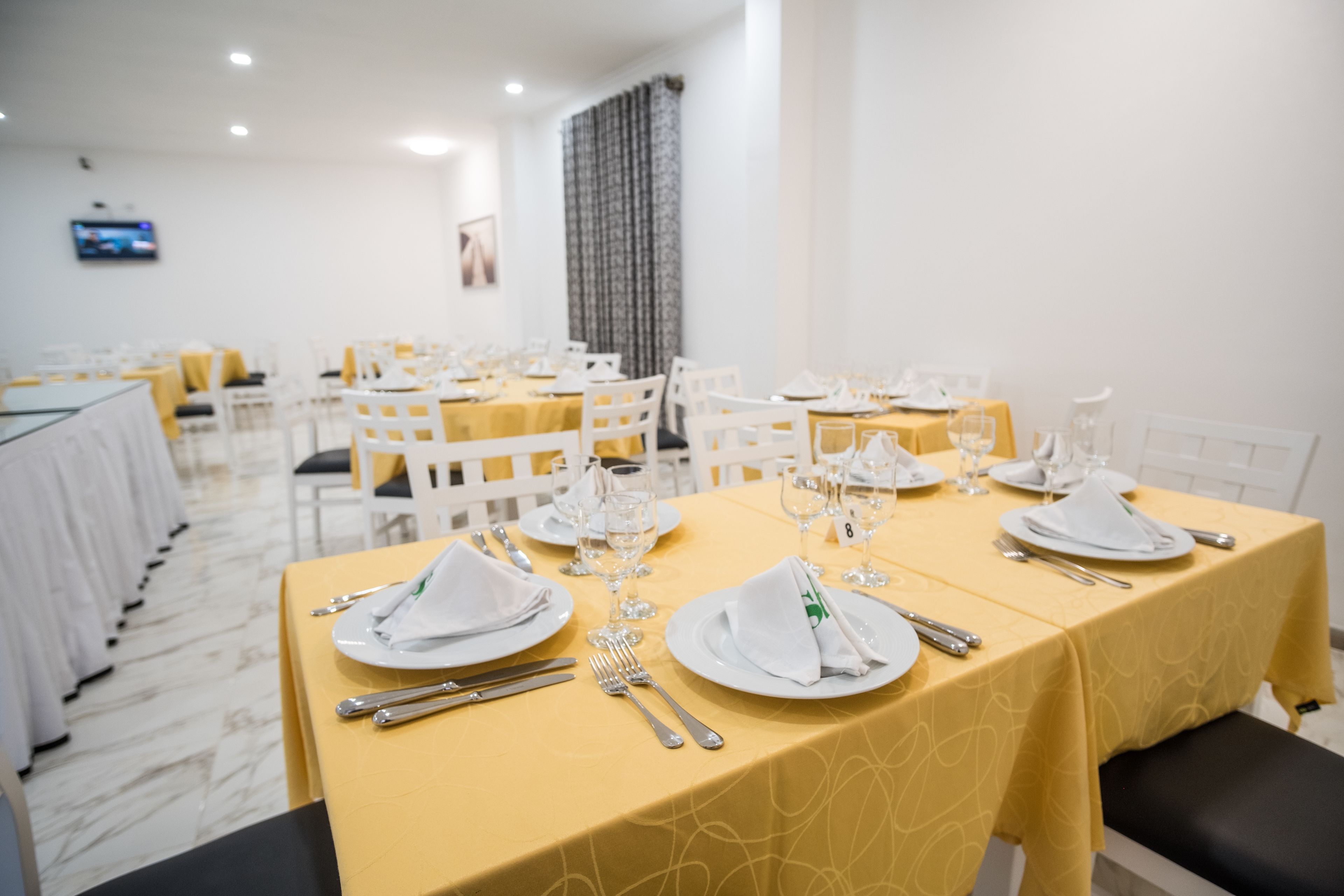 Un comedor con mesas dispuestas para comer, sillas blancas y un televisor en la pared.