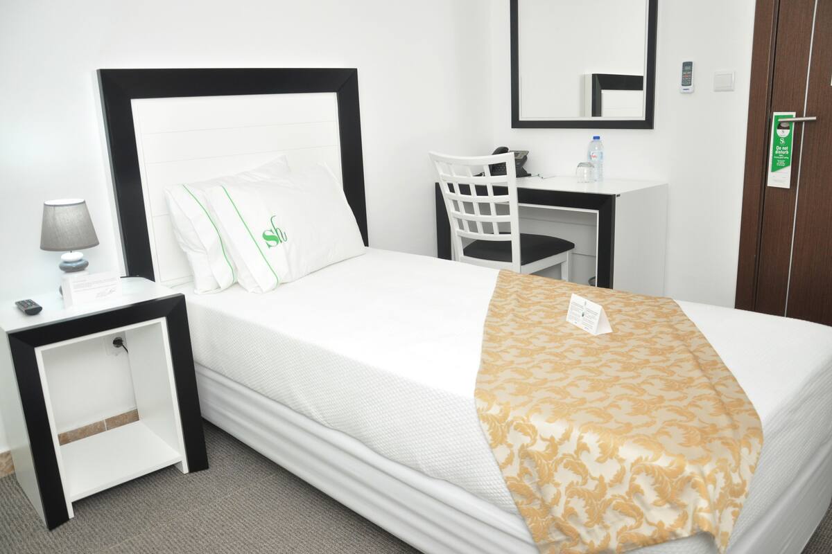 Een keurig opgemaakt bed met een wit en gouden plaid, een nachtkastje met een lamp, een stoel en een bureau met een waterfles.