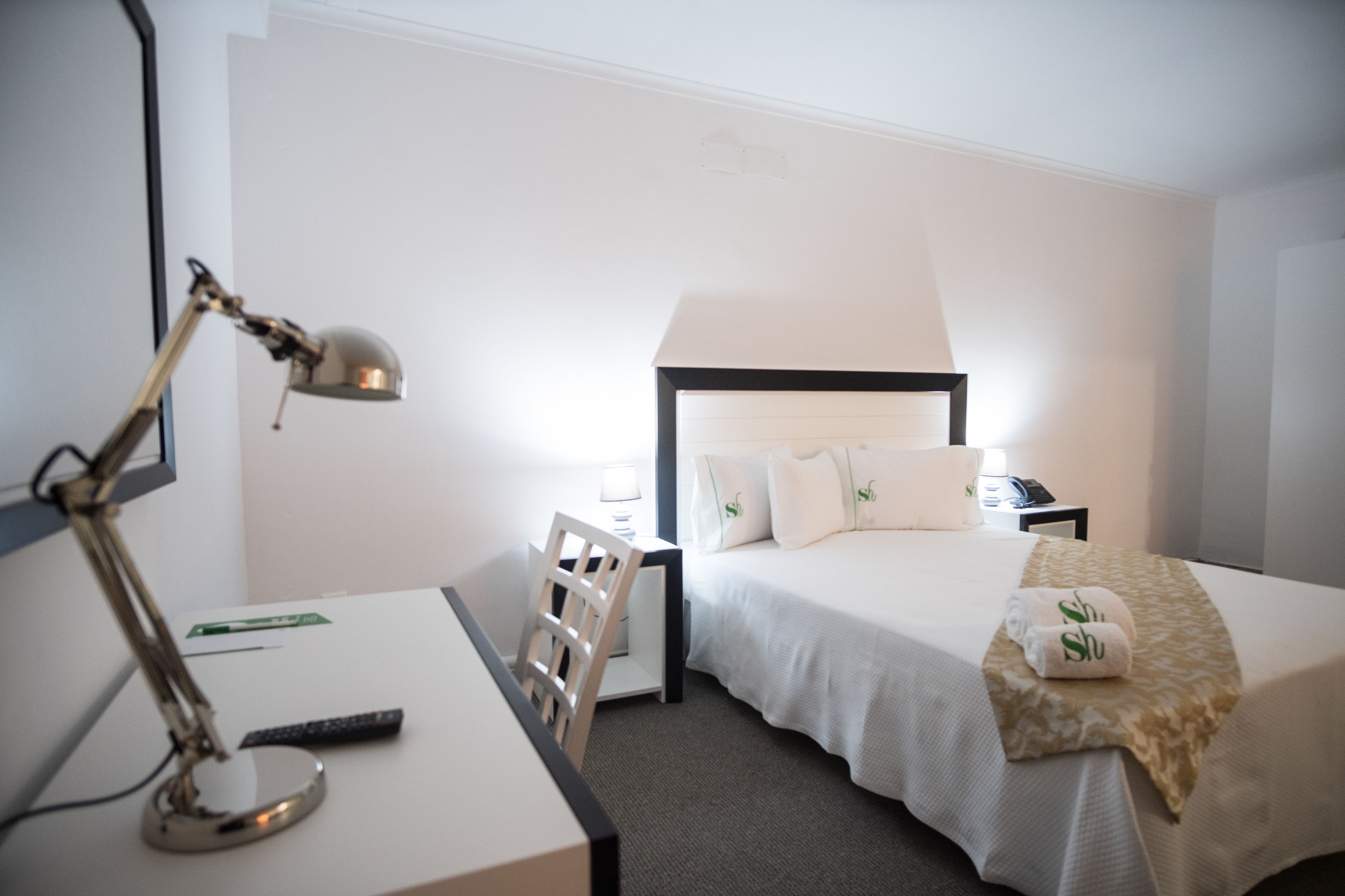 A hotel room with a bed, a desk with a lamp, a chair, and a nightstand with a lamp.