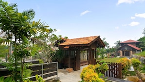 A wooden gazebo with a tiled roof, surrounded by lush greenery and a paved pathway leading to a house with a red roof. - Villa Kiambang (Seremban)