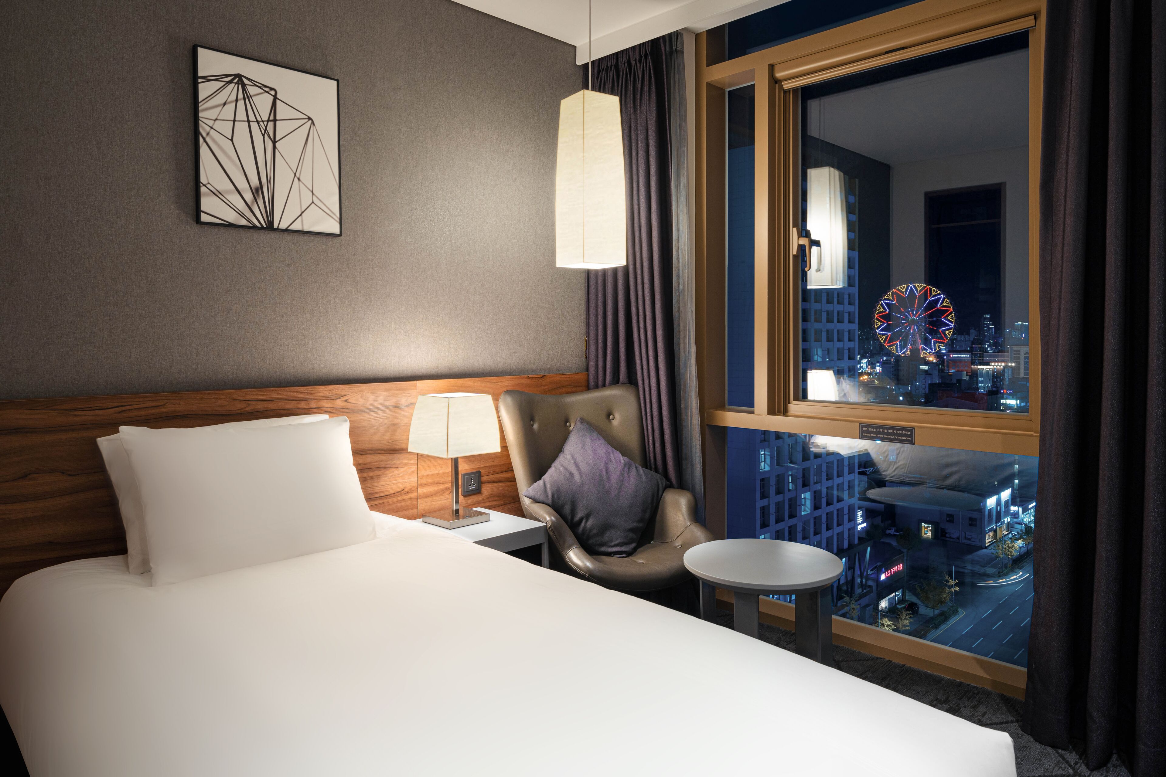 A hotel room with a bed, a chair, a small table, a lamp, and a window with a city view.