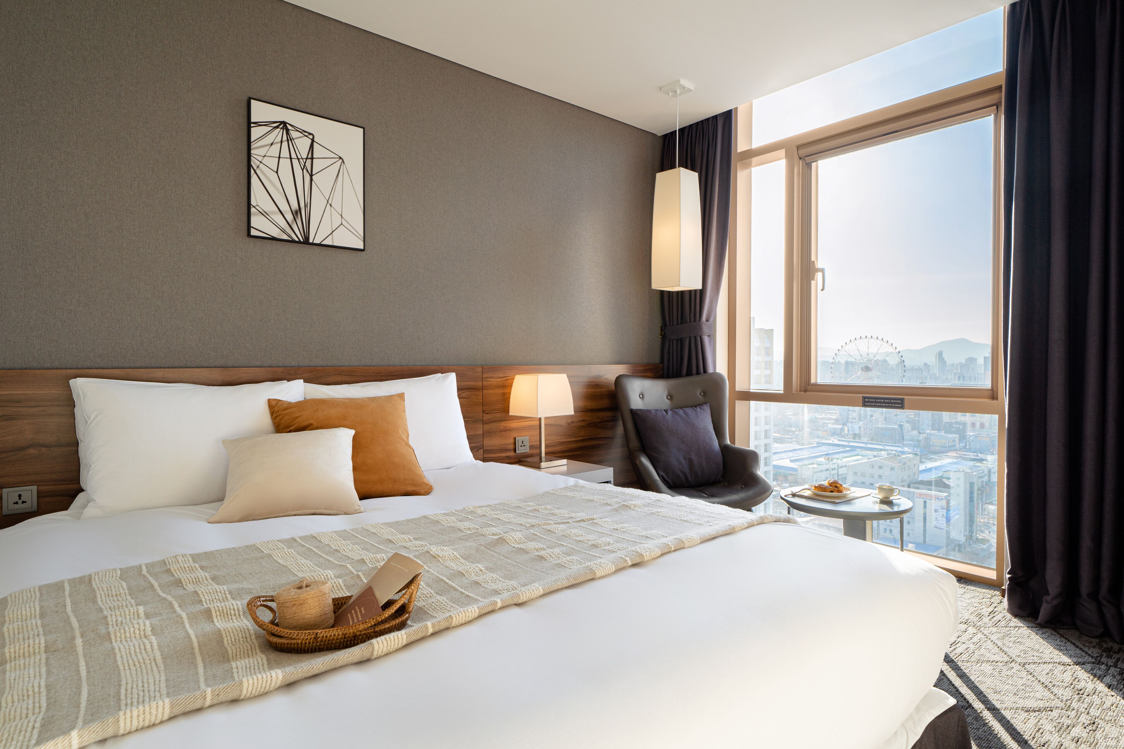 A hotel room with a large bed, a view of the cityscape, a modern lamp, and a chair by the window.