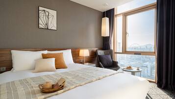 A hotel room with a large bed, a view of the cityscape, a modern lamp, and a chair by the window.