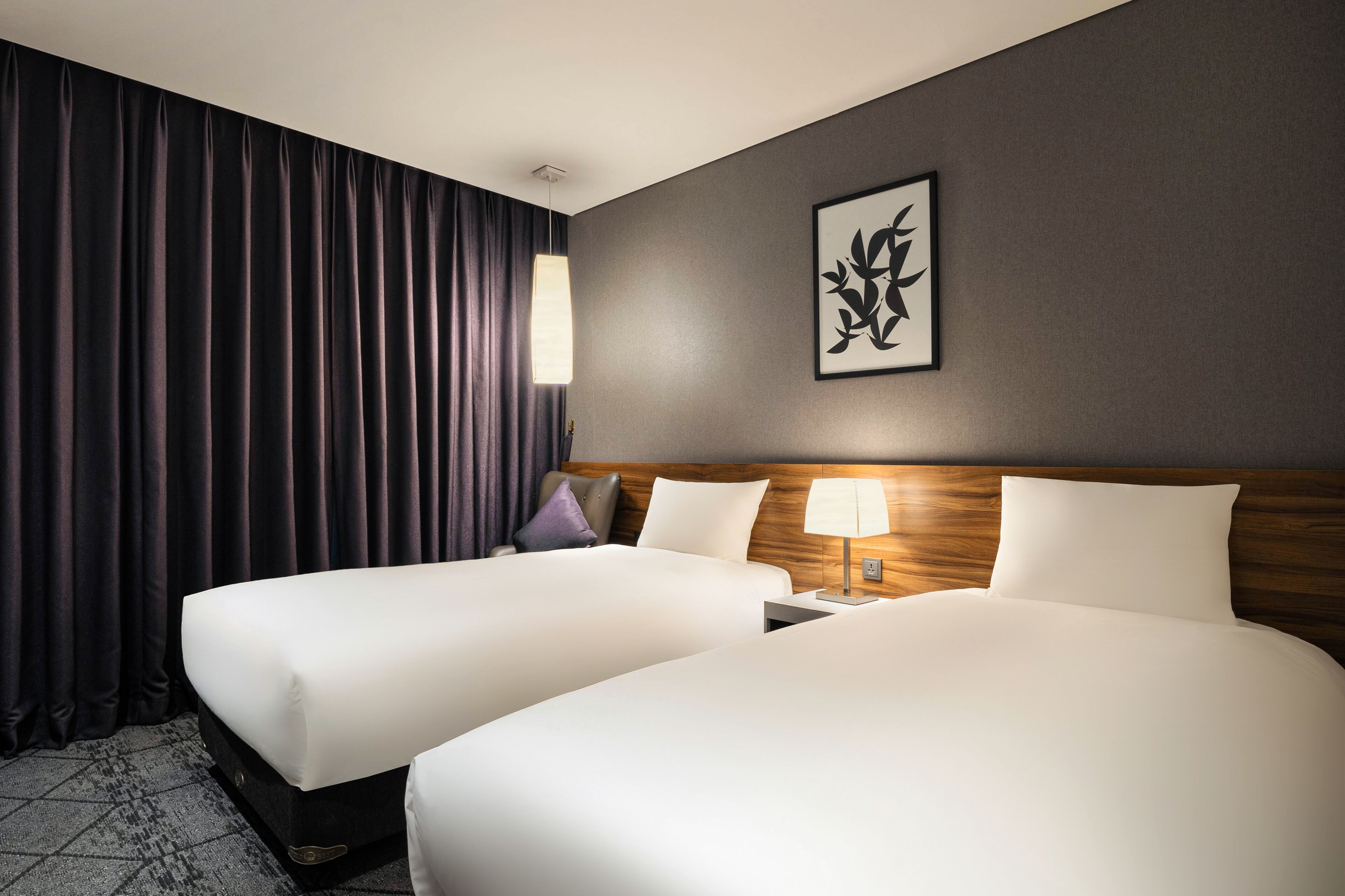 A hotel room with two beds, a nightstand with a lamp, a wall-mounted artwork, and dark curtains.