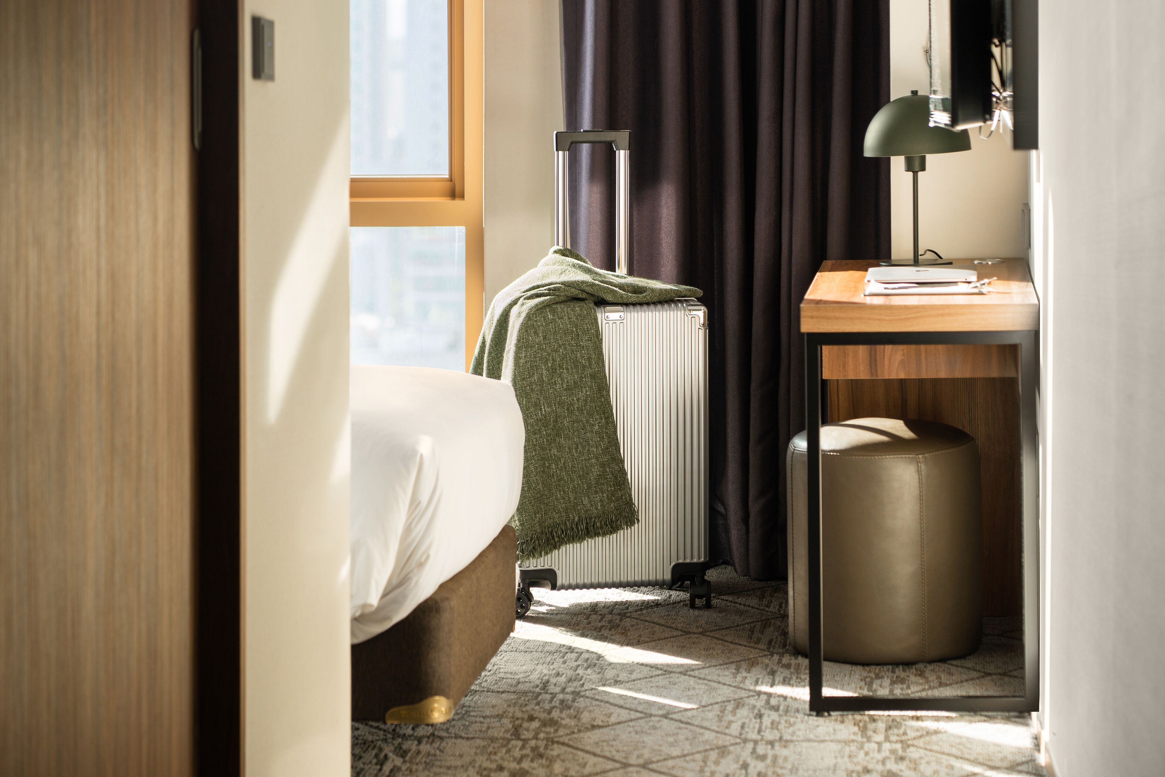 A hotel room with a bed, a radiator, a desk with a lamp, and a suitcase.